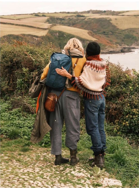 Two women in knitwear embrace on a coastal path, overlooking a grassy, cliffside landscape with a view of the ocean.