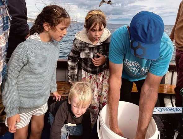 A group of children and an adult fishing off a boat, gathered around a bucket, with the ocean and cloudy sky visible in the background.