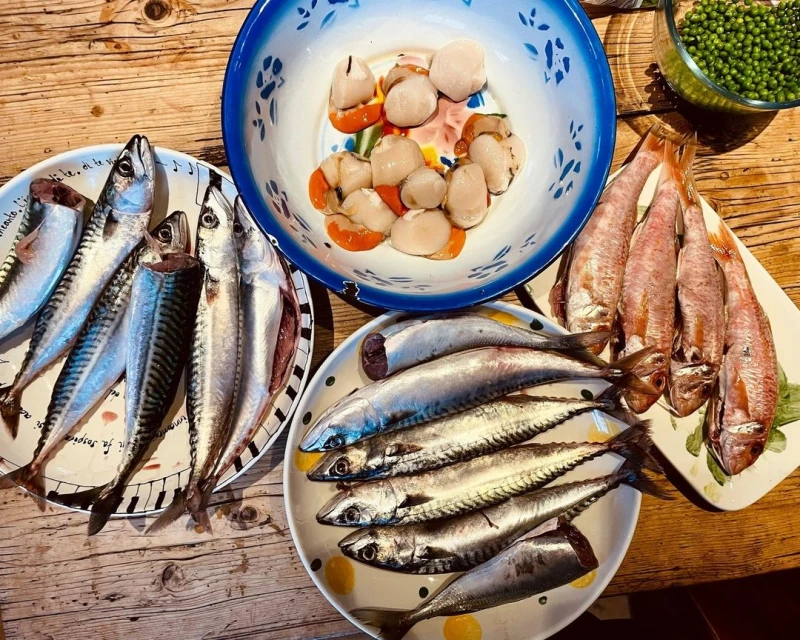 Overhead shot of various types of fresh fish and scallops in bowls and on plates on a wooden table, ready for cooking.