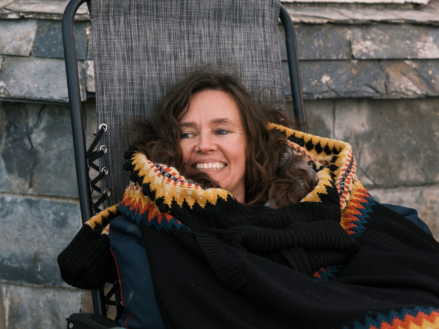 Smiling woman lounges in a chair, wrapped in a colourful blanket on a grey slate background. She appears relaxed and comfortable.