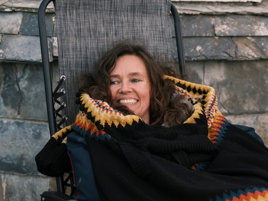 Smiling woman lounges in a chair, wrapped in a colourful blanket on a grey slate background. She appears relaxed and comfortable.