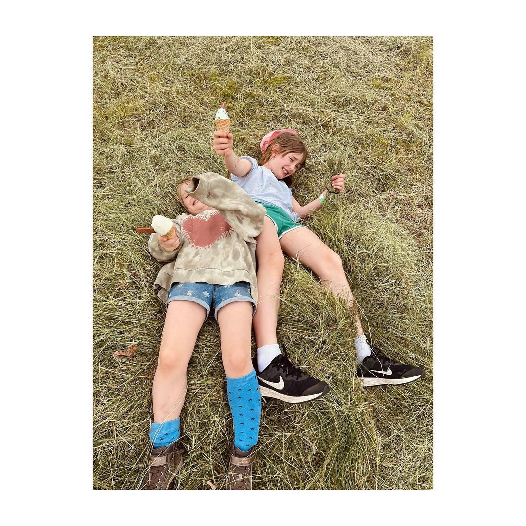 Two young girls lie in a grassy field, holding ice cream cones. One wears denim shorts and blue socks, the other green shorts. 