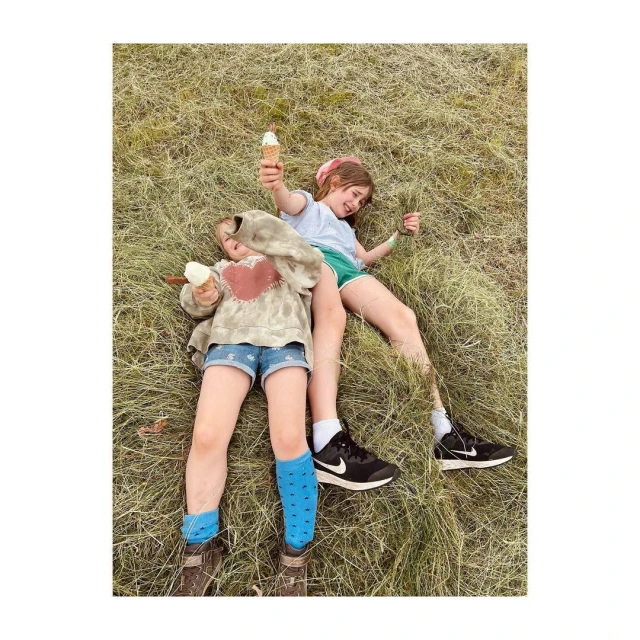 Two young girls lie in a grassy field, holding ice cream cones. One wears denim shorts and blue socks, the other green shorts. 