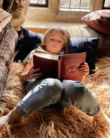 Young child reclining on a plush shaggy rug and dark pillows while reading an open vintage book inside.