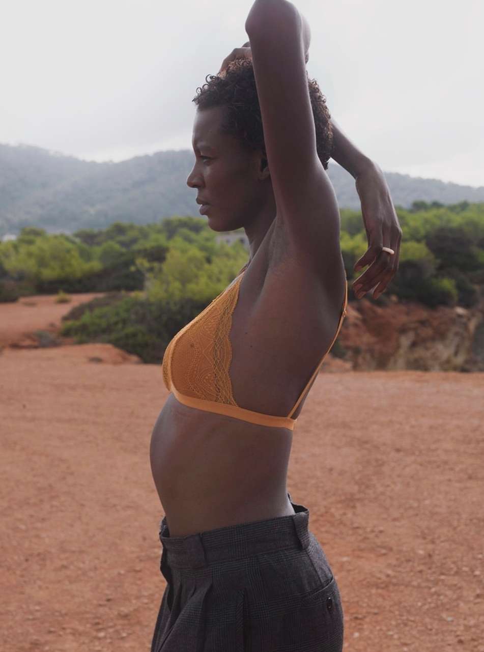 Side profile of a woman in a yellow bra and grey pants in a landscape with mountains and green trees under an overcast sky.
