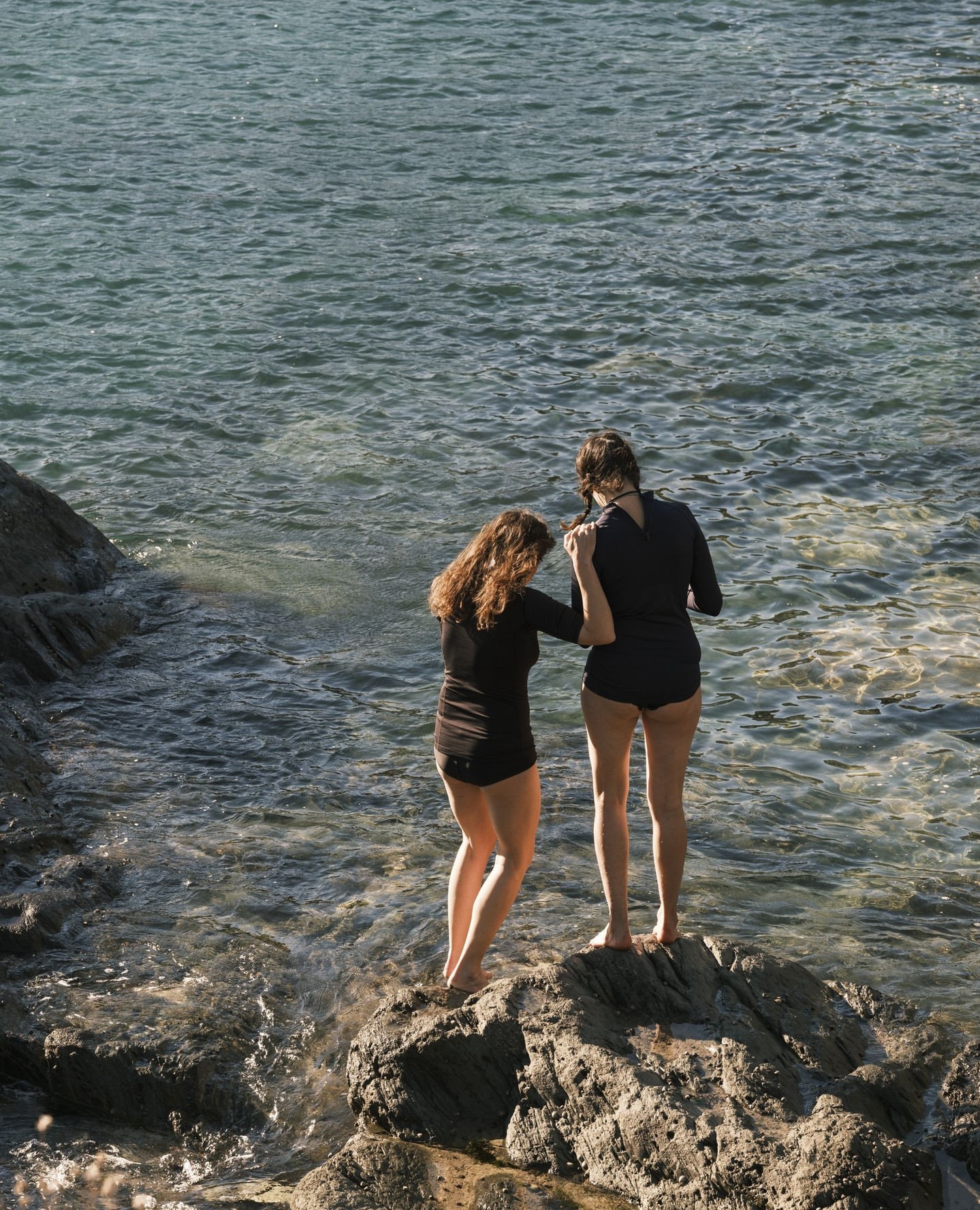 Two women in swimwear stand on rocks at the edge of a sea. The women face the water, one with hand on the other's arm.