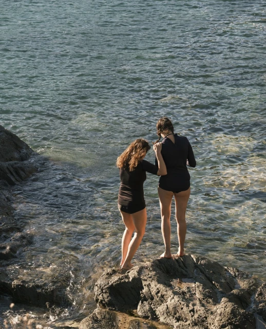Two women in swimwear stand on rocks at the edge of a sea. The women face the water, one with hand on the other's arm.