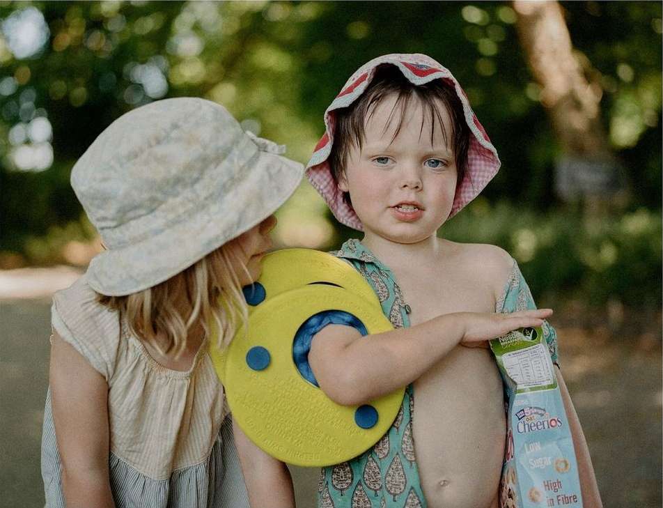 Two young children in sun hats stand in a sun-dappled park with one child holding a box of Cheerios cereal and a yellow kickboard.