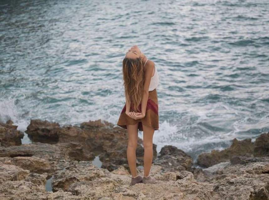 Adult woman with long brown hair standing on rocky beach with ocean waves crashing in the background on a cloudy day.