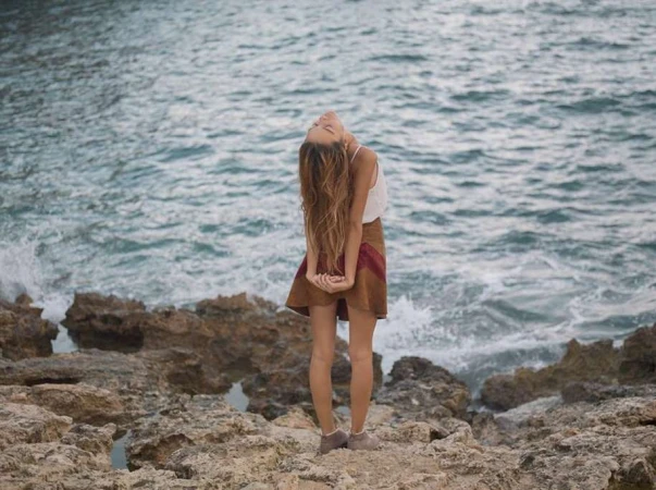 Adult woman with long brown hair standing on rocky beach with ocean waves crashing in the background on a cloudy day.