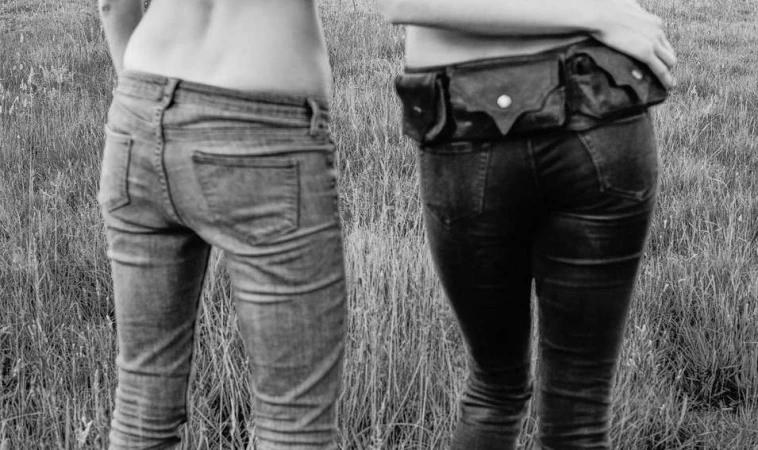 Monochrome shot of two women in jeans standing in a field of grass, one wearing a waist bag, showcasing fashion and outdoor style.