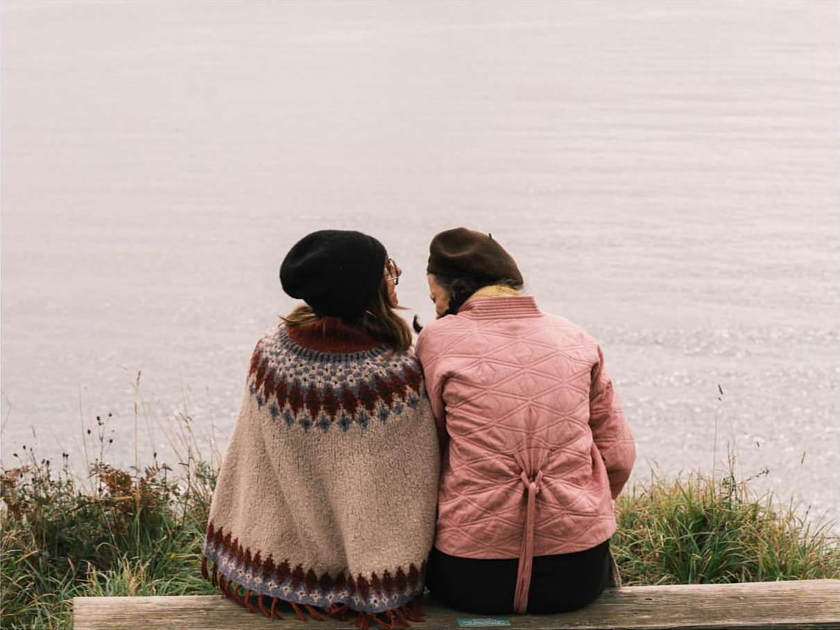 Two women sit on a wooden bench overlooking a calm body of water, wearing cozy outerwear like sweaters and jackets in an outdoor setting.