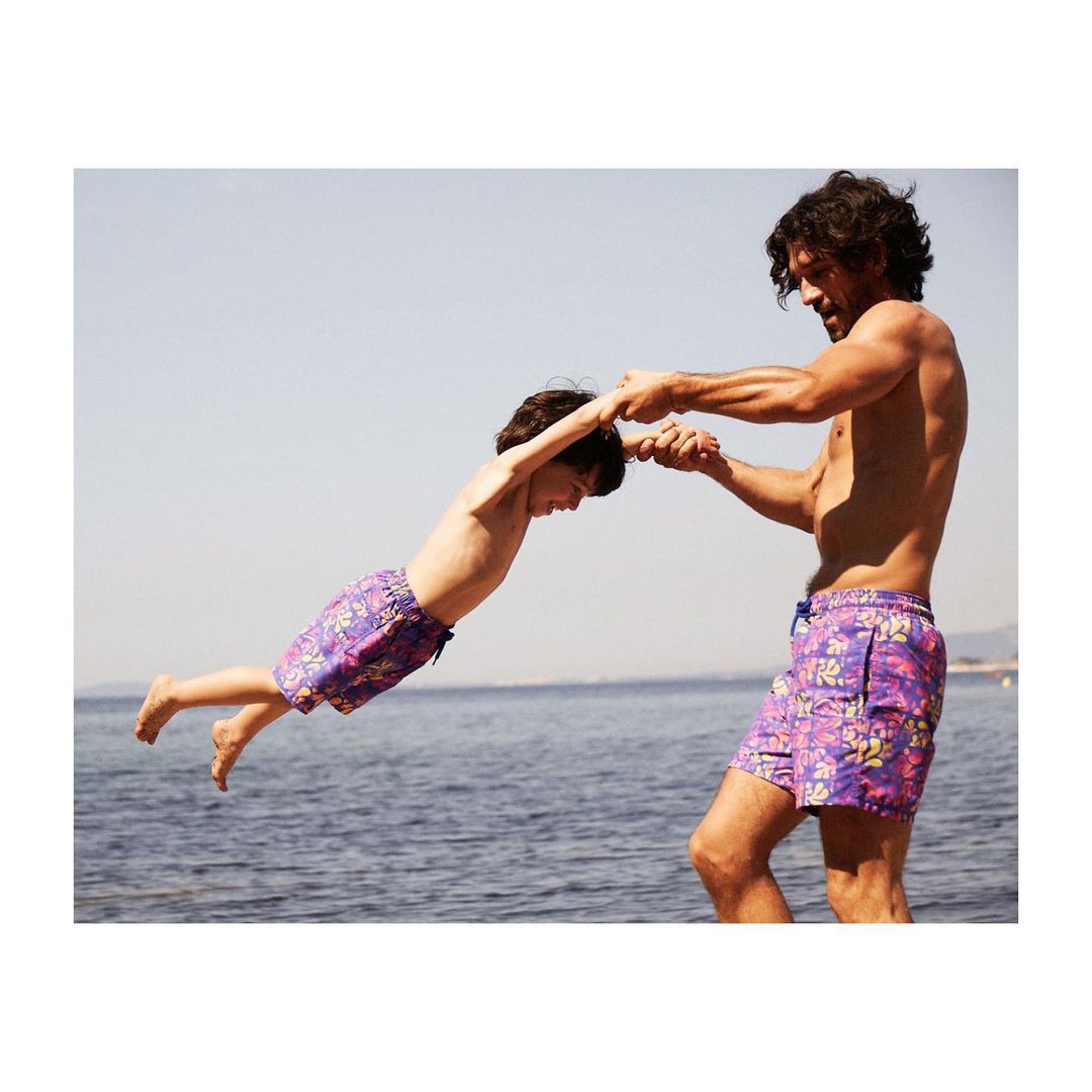 Father and son play at the beach; the shirtless dad swings his son, who wears floral swim trunks, with the ocean in the background.