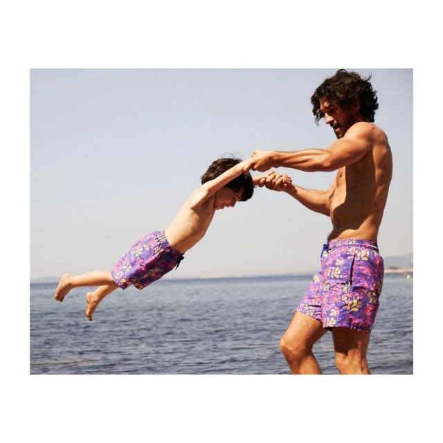 Father and son play at the beach; the shirtless dad swings his son, who wears floral swim trunks, with the ocean in the background.