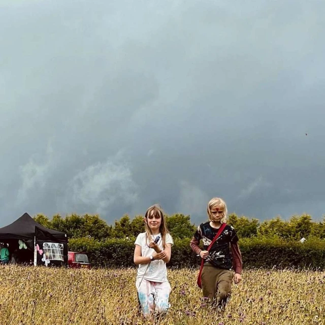 Two young girls walk through a field of wildflowers under an overcast sky, with a black tent and a vehicle in the background.