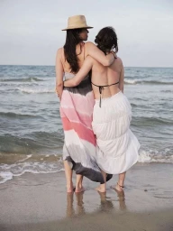 Two women with long dark hair embracing on a sandy beach looking out towards the calm ocean water