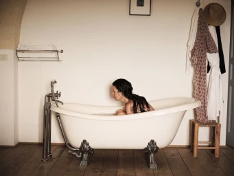Woman with long dark wet hair sitting inside a white clawfoot bathtub in a rustic bathroom.