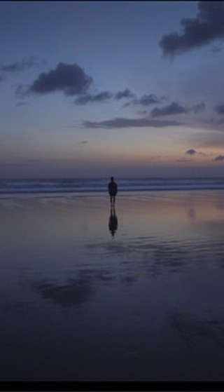 Person stands on the reflective shore during twilight, as soft waves reach the beach under a gradient sky of blues and oranges.