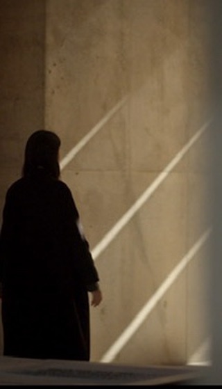 A woman in a black coat walks near a textured wall with light streaks, creating a contrast of dark clothing against the bright background.