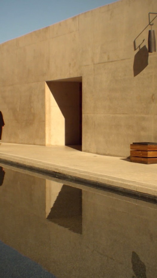 Modern architecture featuring a tan-colored wall with an open doorway, complemented by a tranquil pool reflecting the structure's design.