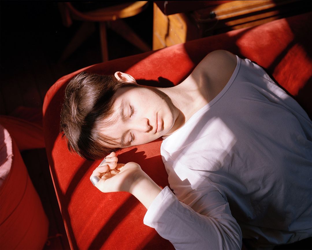 Woman sleeping on a red velvet couch bathed in harsh sunlight creating shadows, wearing a white top in an indoor environment.