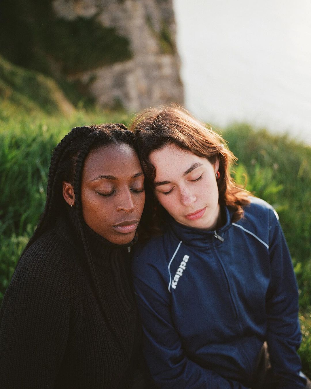 Close-up of an African-American woman and a Caucasian woman with eyes closed, heads touching, wearing casual clothes in a grassy, outdoor setting.