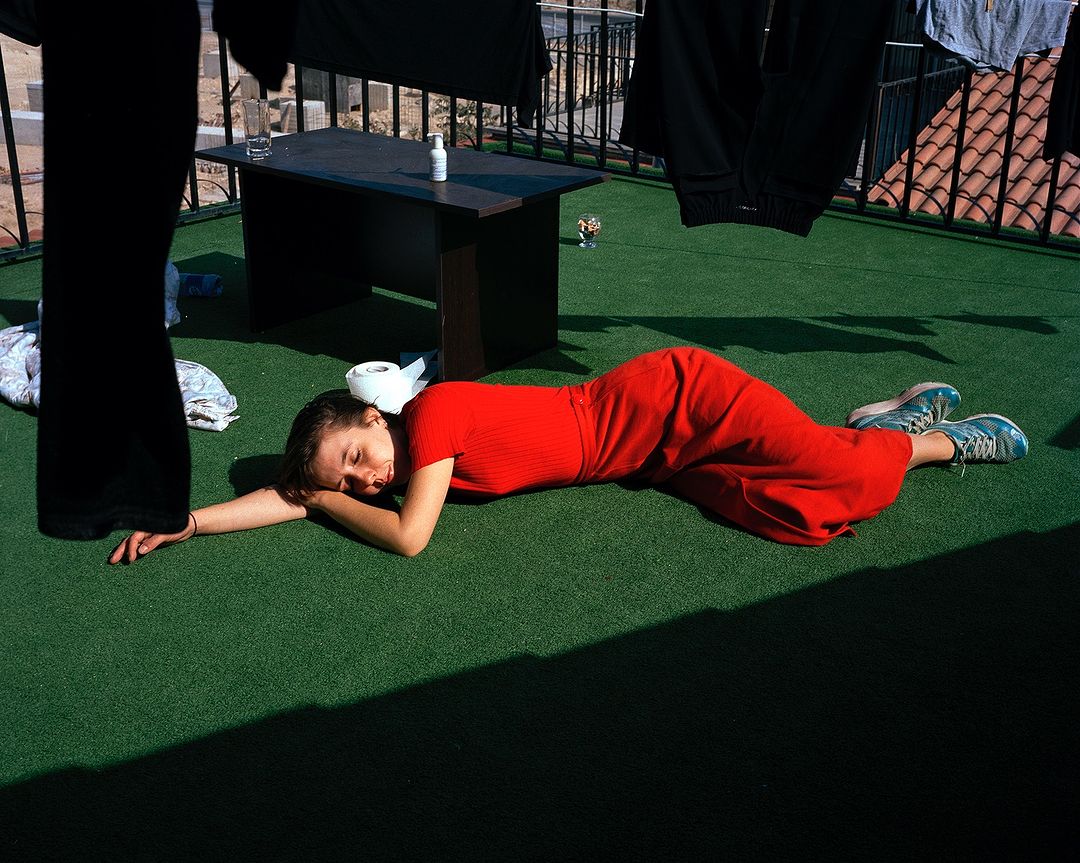 Woman in red dress resting on green astroturf next to black table with personal care items and drying clothes in the background.