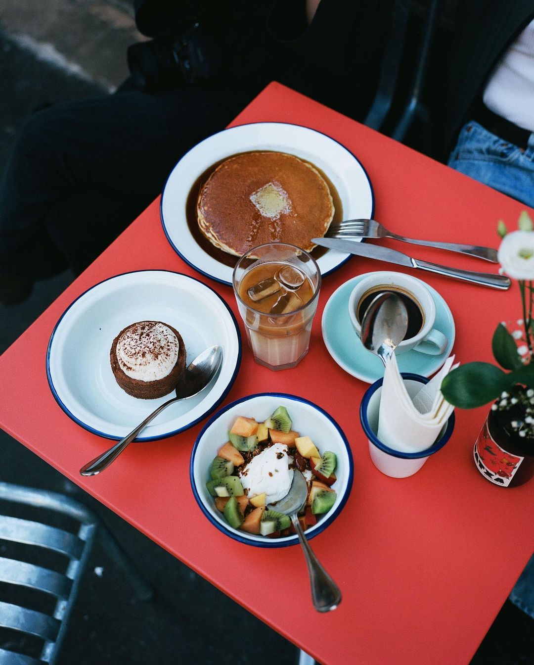 Overhead shot of a red table featuring pancakes, fruit salad, coffee, iced latte, and dessert, served in white enamelware dishes.