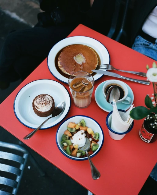 Overhead shot of a red table featuring pancakes, fruit salad, coffee, iced latte, and dessert, served in white enamelware dishes.