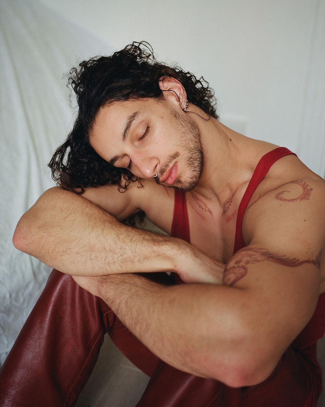 Young adult man with curly hair and tattoos sits indoors on white fabric in red tank top and pants, eyes closed, resting in studio.