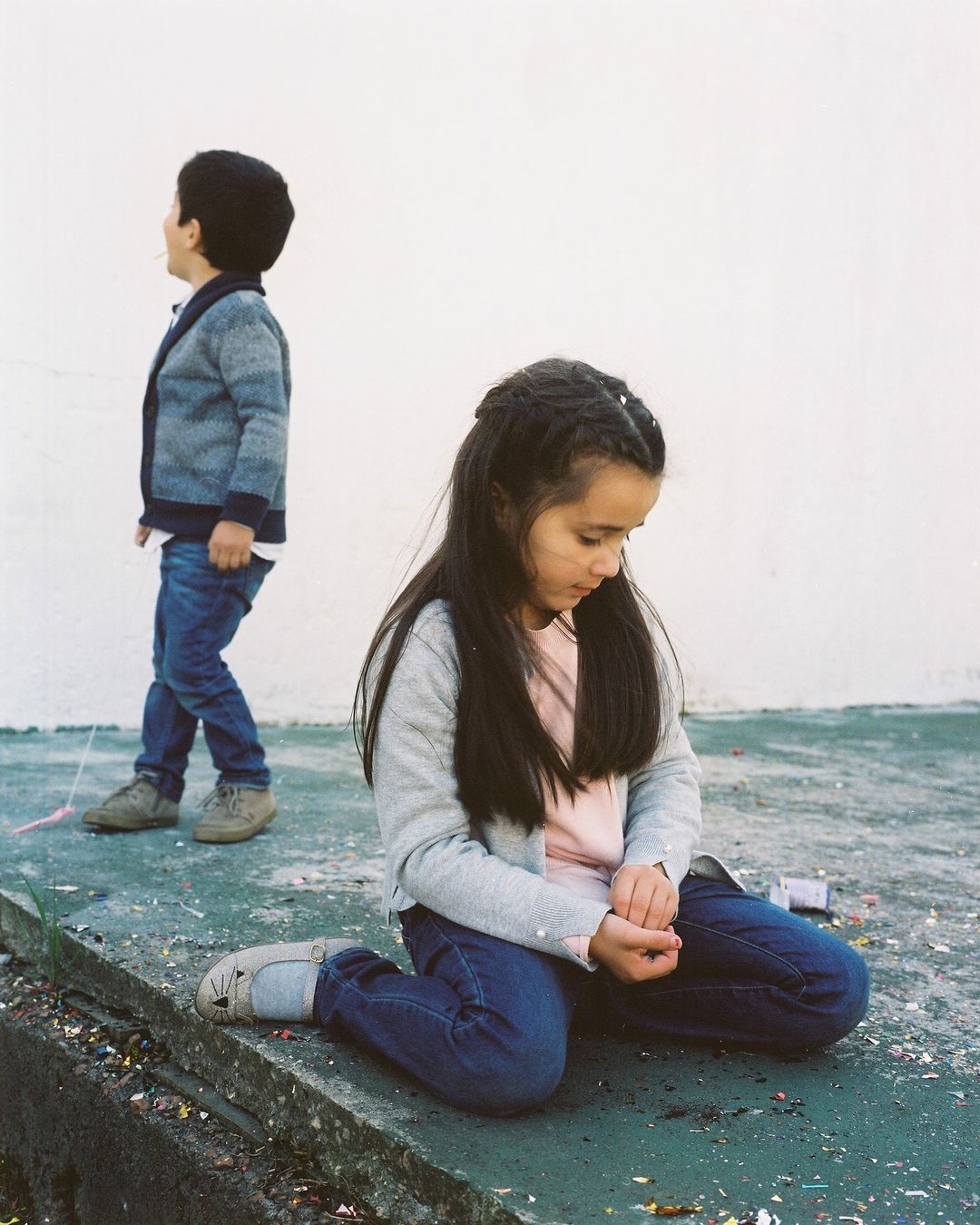 Two children in outdoor setting. A girl kneels with head down while a boy walks behind her. They are wearing casual clothing.