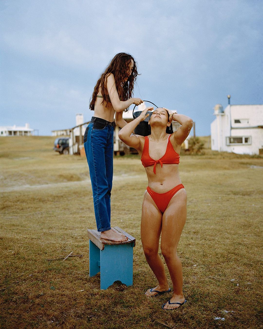 Two women in swimwear outdoors; one stands on a stool pouring water from a black bucket over the other's head on a grassy landscape.
