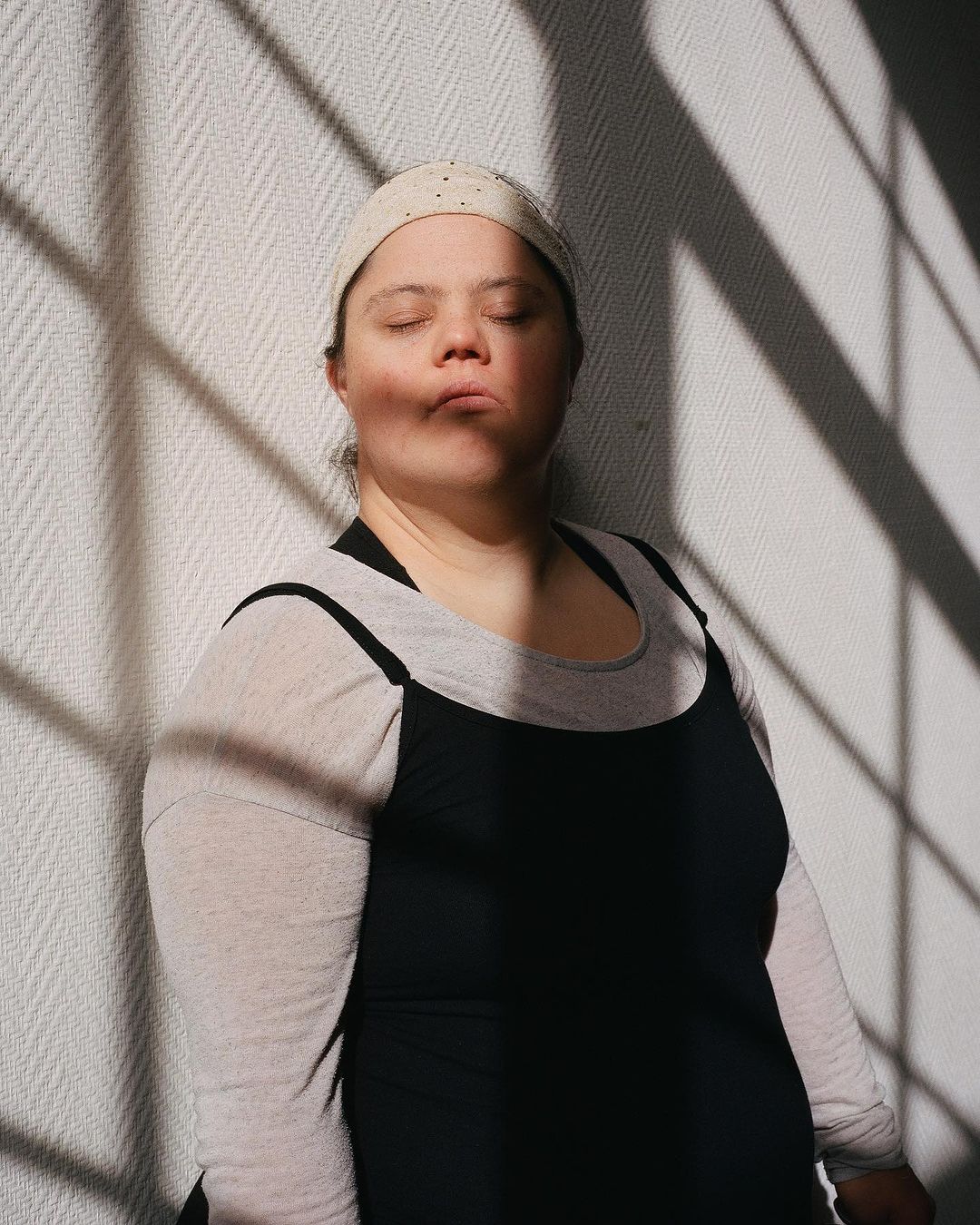 Portrait of a woman with Down syndrome standing in front of a textured wall with strong shadow lines from a window, wearing casual clothing.