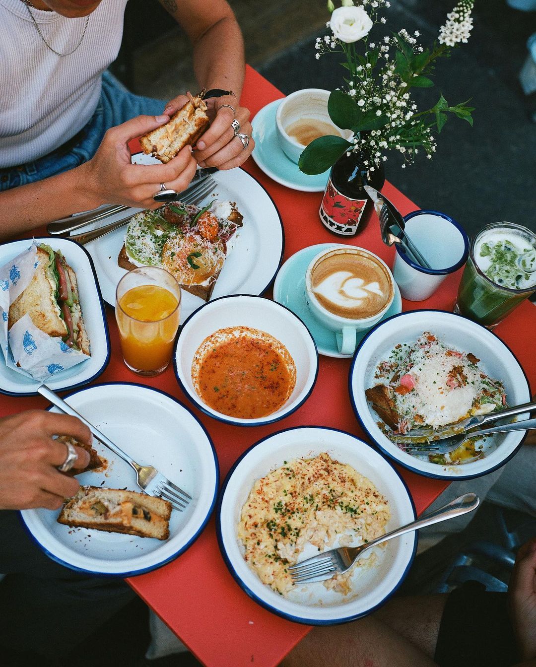 Overhead shot of people dining outdoors at a cafe with a variety of sandwiches, egg dishes, juice, and coffee on a red table.