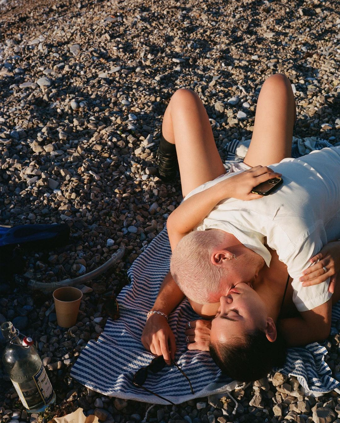 Intimate moment of a couple embracing and kissing on a rocky beach, bathed in warm sunlight, evoking a sense of natural intimacy.