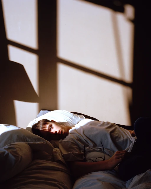 Indoor portrait of a young adult lying in bed in front of a window with sunlight creating shadows on the white wall behind.