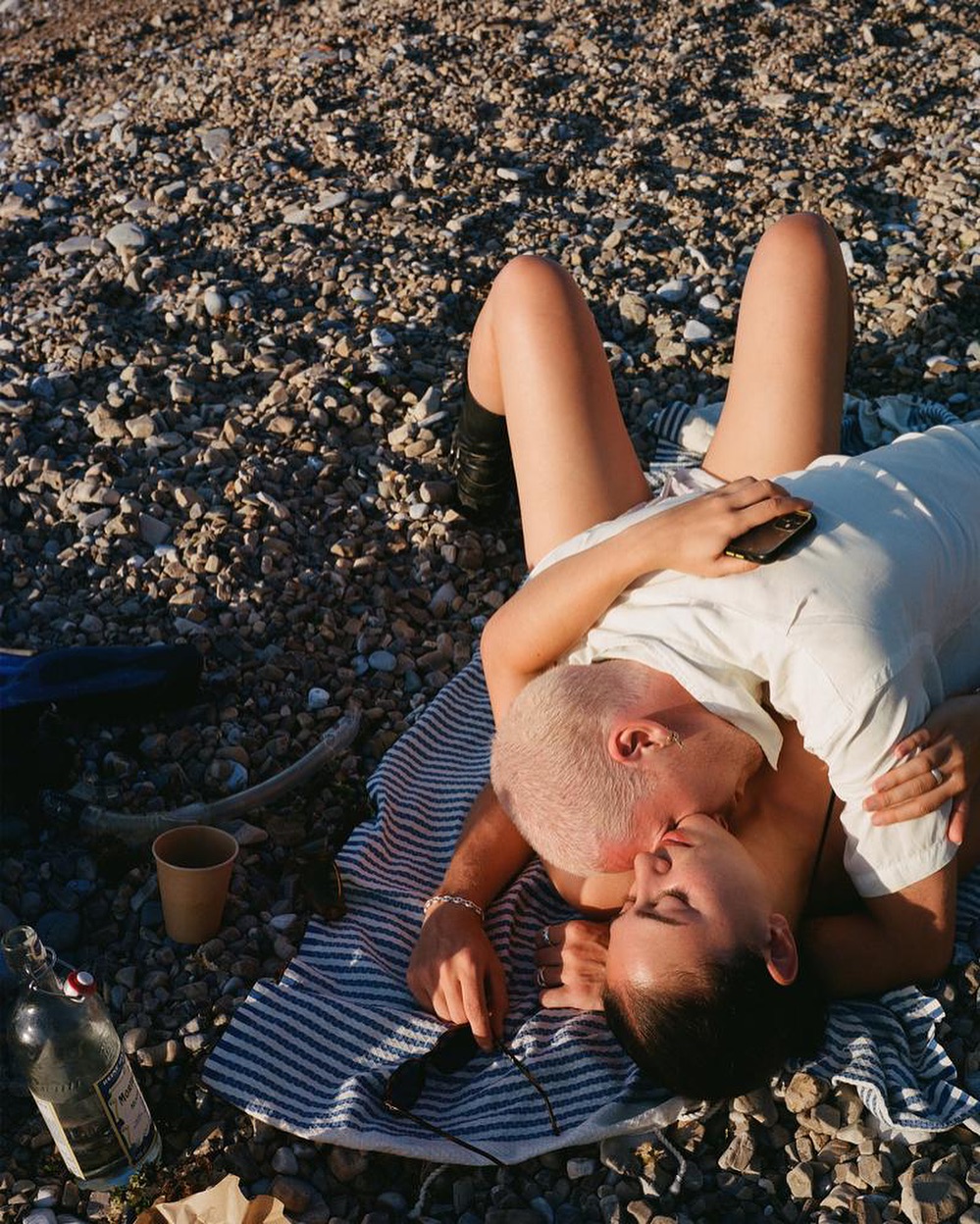 Couple kissing while lying on a striped blanket on a rocky beach with a beverage and a bottle nearby, captured in natural light.