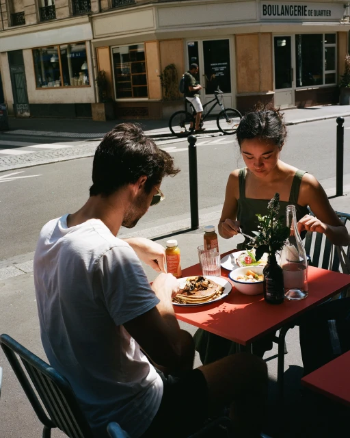 A young couple dining outdoors at a cafe on a sunny day in an urban setting. They are eating food and drinking beverages at a red table.