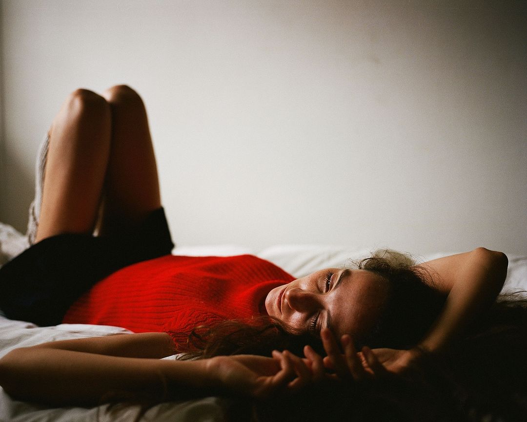 Woman in red top and black shorts lying on a white bed with her legs raised against a white wall in a relaxed pose.