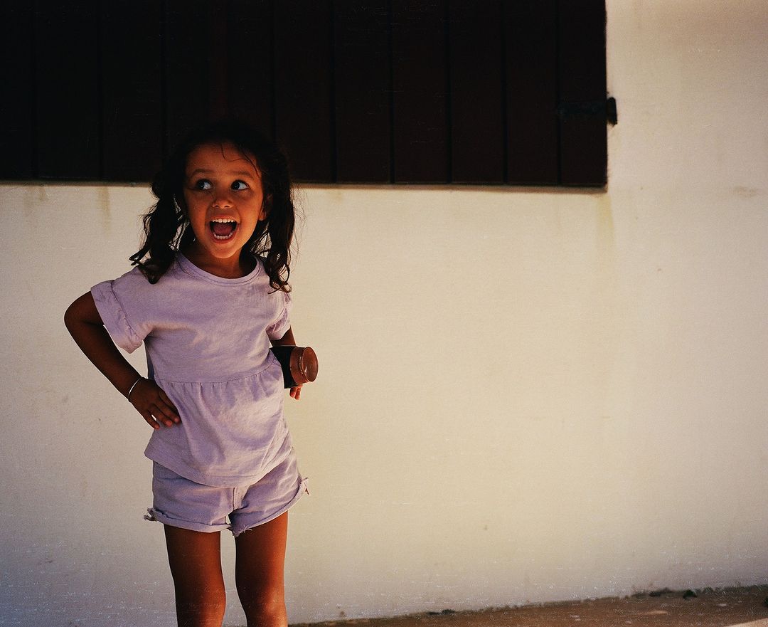A cheerful young girl with dark hair and wearing a matching lilac top and shorts stands against a textured white wall with an open mouth.