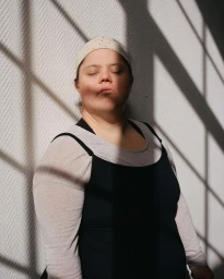 Portrait of a woman with eyes closed wearing a black dress and white headband under harsh window light shadows by Vera Prego.