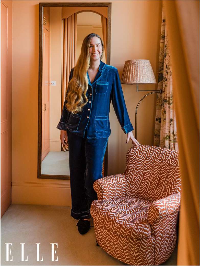 Adult woman in blue velvet pajamas standing in a room with a peach-colored wall, next to an armchair, and a mirror.