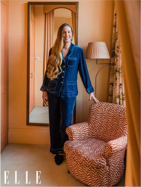 Adult woman in blue velvet pajamas standing in a room with a peach-colored wall, next to an armchair, and a mirror.