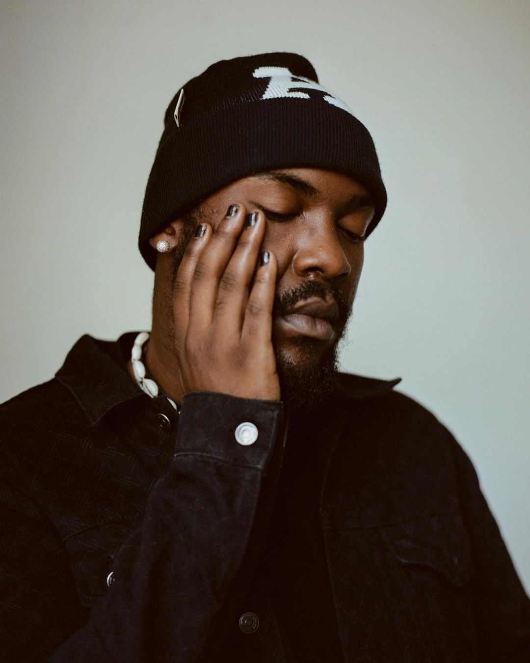Close-up of an adult Black man with his eyes closed, wearing a black beanie and black jacket, set against a light gray background.