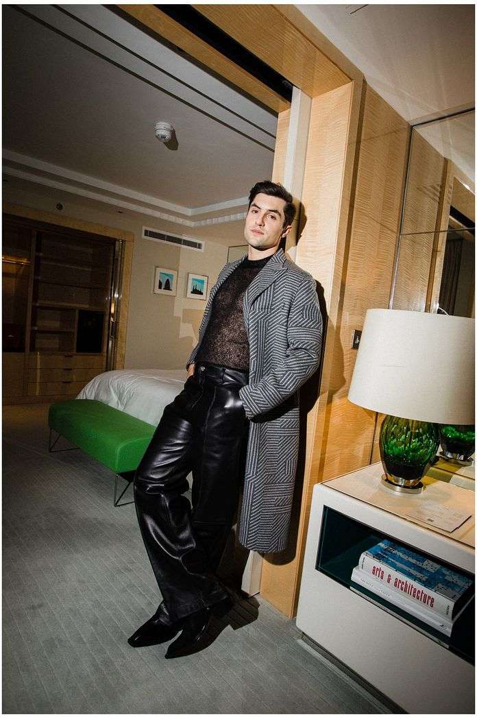 Man poses indoors wearing a grey and black striped coat, black mesh top, shiny black pants, and pointed shoes, leaning against a column.