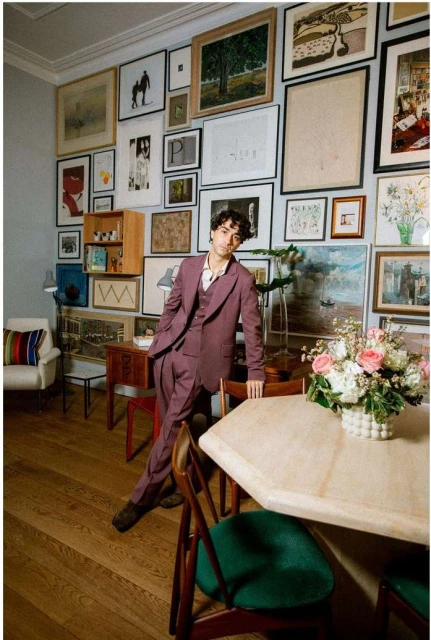 Man in burgundy suit poses indoors with wood floor, octagonal table, and wall of art in various frames, creating a sophisticated ambiance.