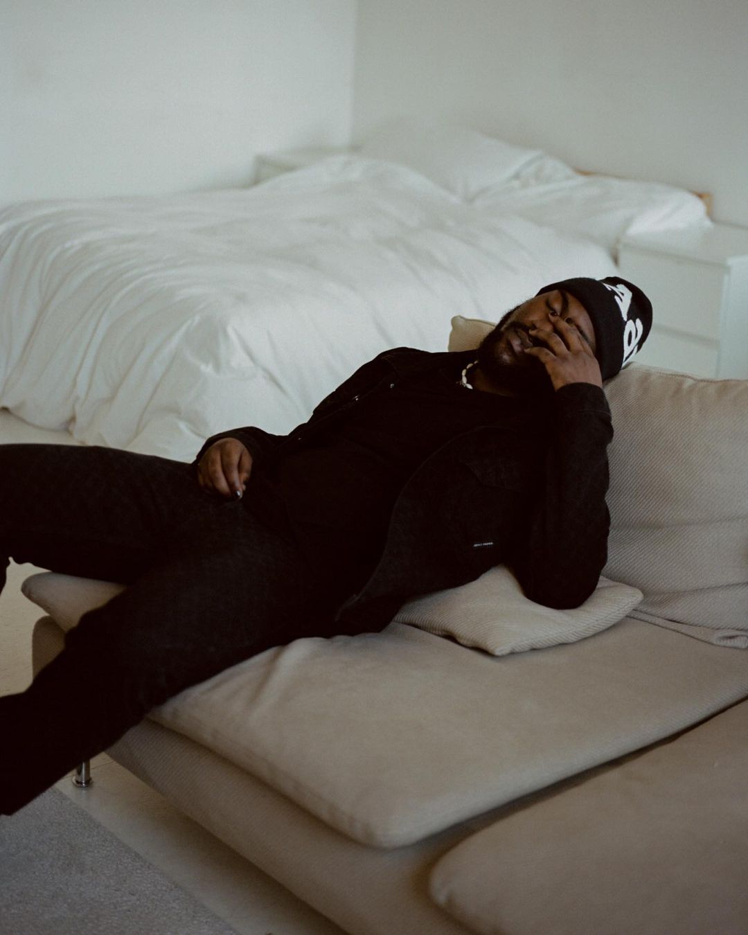 Adult male in black loungewear and beanie relaxing on beige couch in a bedroom setting with white walls, bedding, and minimal decor.