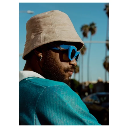 Close-up portrait of a Black man wearing a beige bucket hat and blue sunglasses outdoors against palm trees.