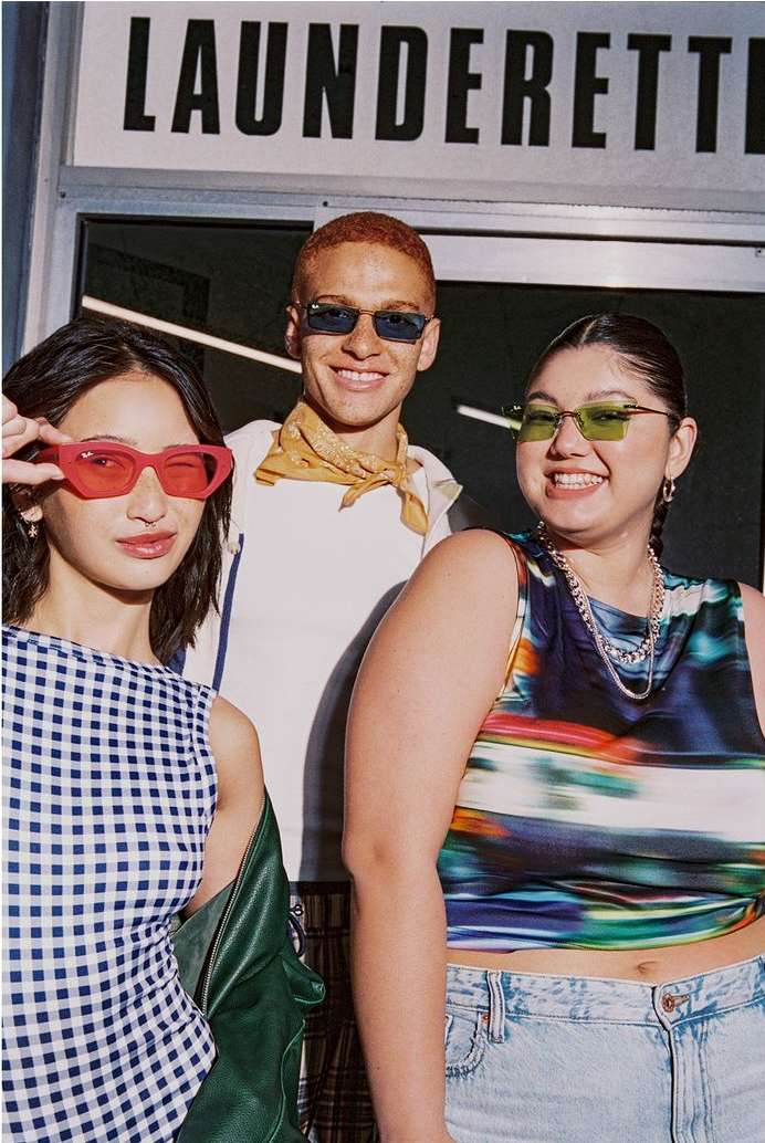 Three fashionable young adults pose outside a laundrette, wearing colorful sunglasses and stylish clothing. They are smiling at the camera.