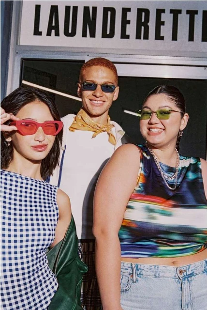 Three fashionable young adults pose outside a laundrette, wearing colorful sunglasses and stylish clothing. They are smiling at the camera.